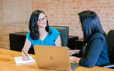 Two women sitting at a table, one taking notes and the other on a laptop.