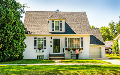 The exterior of a suburban home with someone outside watering the plants at the front windows.