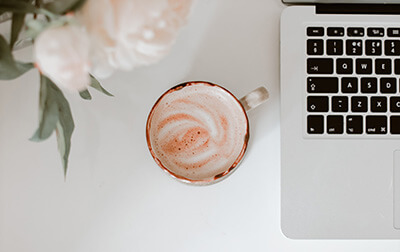 A cup of coffee on a table with a laptop and flowers.